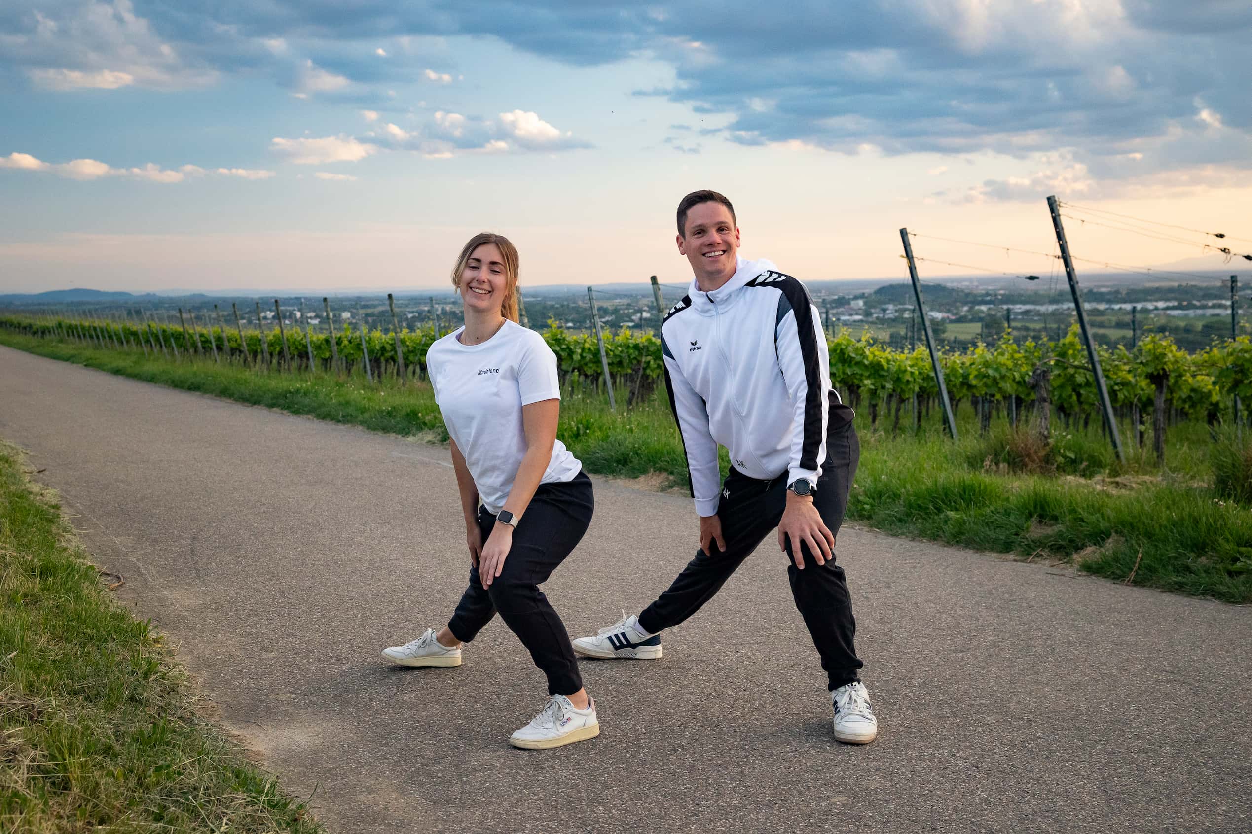 Zwei Vereinsmitglieder in weißen TV-Shirts beim Dehnen auf einem Weg durch die Weinberge